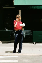 Quinn Mora in sunglasses, a red cropped knit cardigan, and dark wide-leg trousers eating outside a cafe with green awnings.