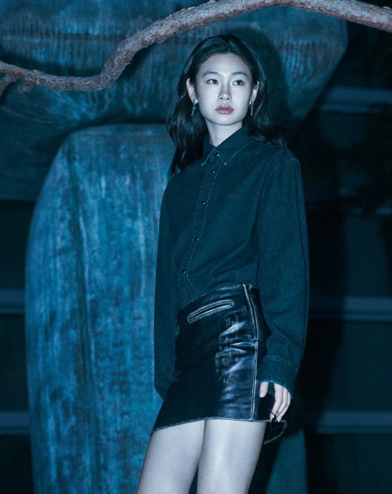 Young woman in a dark denim shirt and black leather miniskirt poses in front of a large blue stone sculpture.