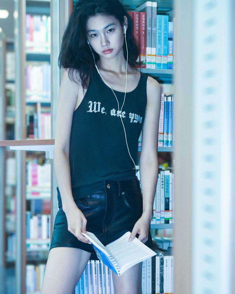 An Asian woman stands in a library wearing a tank top and shorts, holding an open book, with shelves full of books in the background.