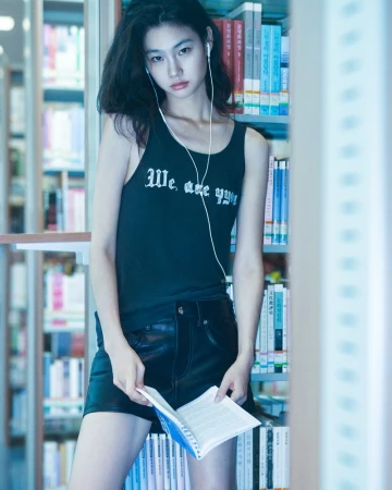 An Asian woman stands in a library wearing a tank top and shorts, holding an open book, with shelves full of books in the background.