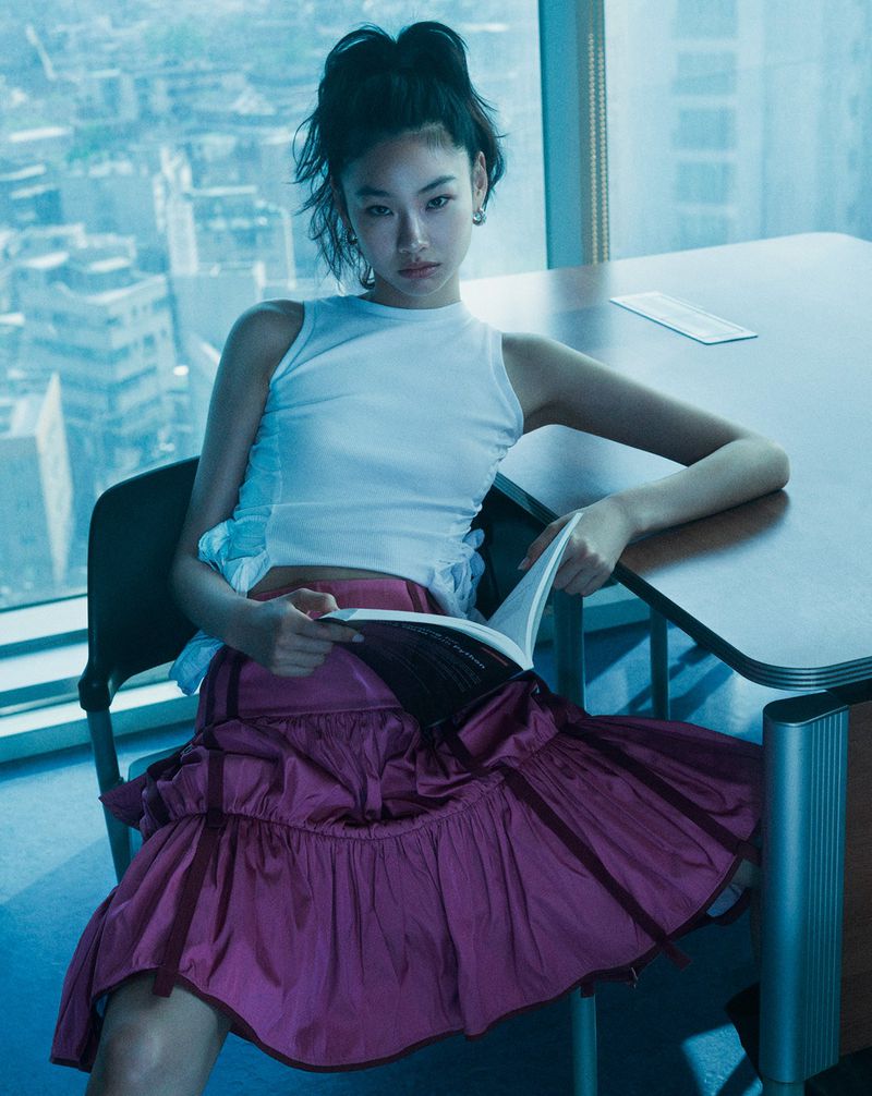 Asian woman in white top and magenta ruffle skirt sits at a desk with a book in a high-rise, bathed in cool, natural light.