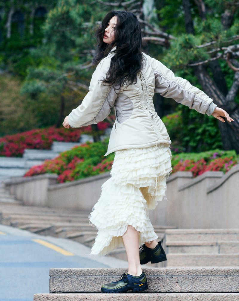 An adult female model poses on outdoor steps in a cream-colored jacket and ruffled skirt, with trees and foliage in the background.
