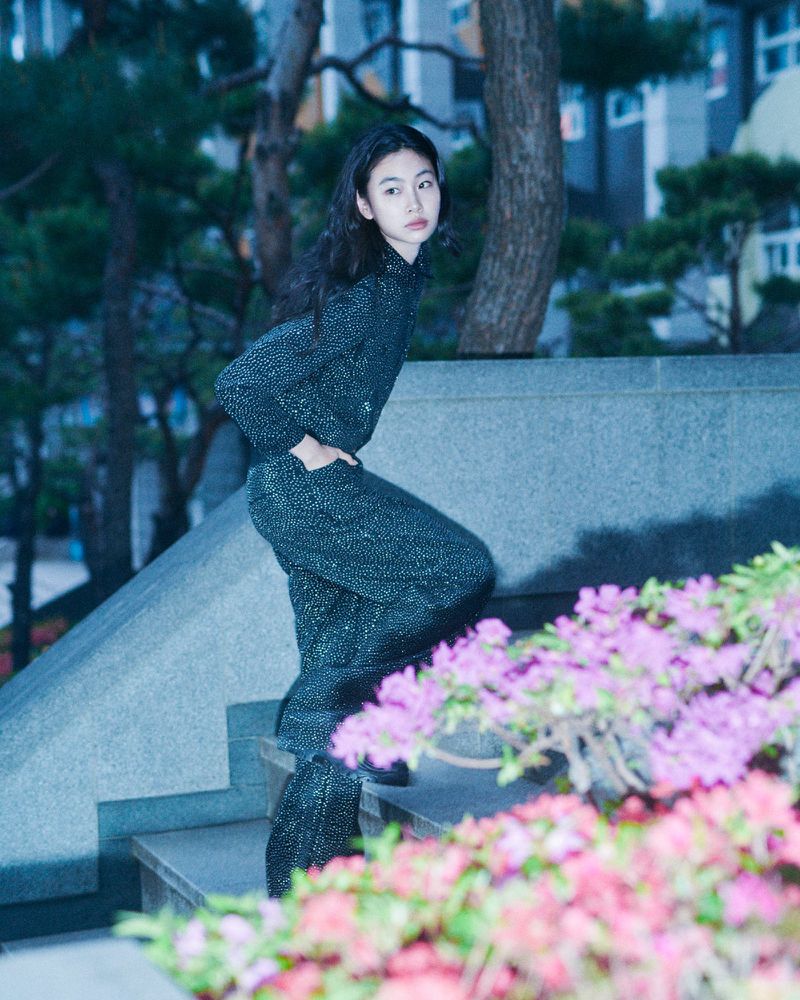 Asian woman in sparkling outfit poses on stairs outdoors with blooming flowers and blurred buildings in the background during daytime.
