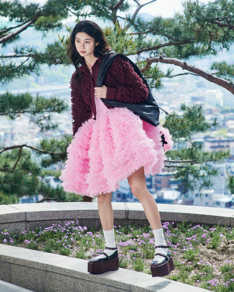 Hoyeon Jung standing on a stone ledge in a voluminous pink tulle dress, burgundy textured jacket, and platform shoes.