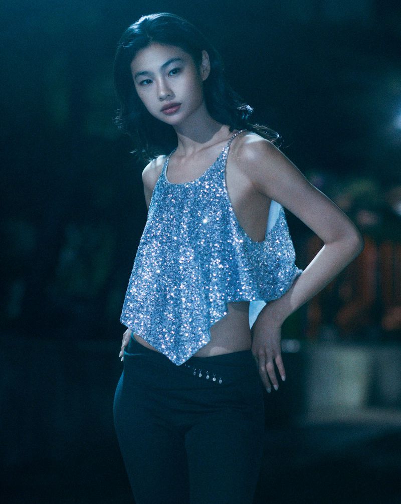 Young woman in shimmering silver crop top and dark pants poses against a blurred background, captured in a medium shot.