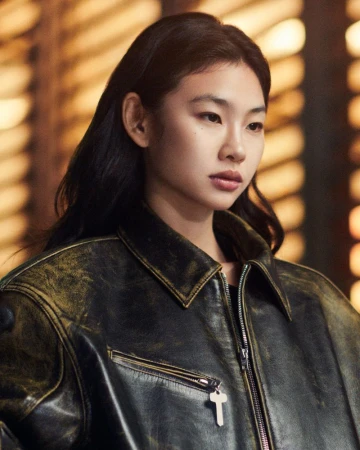 Hoyeon Jung wearing an oversized distressed black leather jacket with a key zipper pull against a warm bokeh background