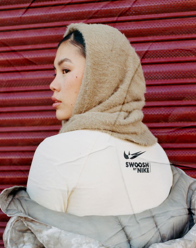 Woman with tan, hooded scarf and beige shirt featuring the Nike swoosh logo in front of a corrugated red metal wall.