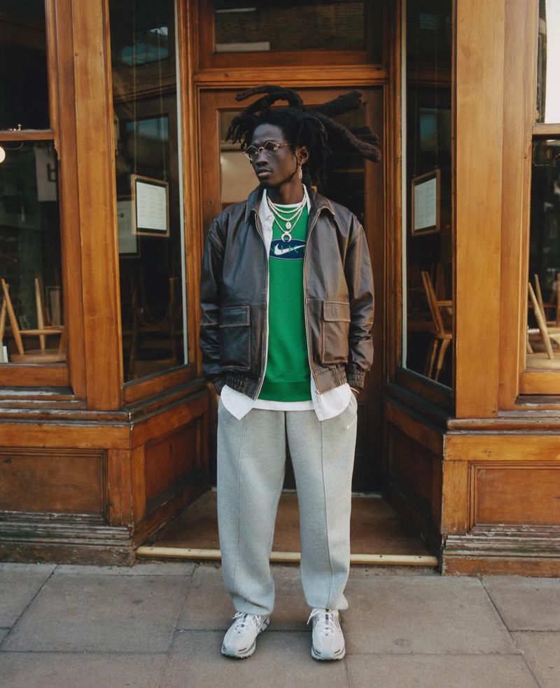 A Black male model stands outside a wooden-framed building, wearing a leather jacket, green Nike sweatshirt, grey pants, and white sneakers.