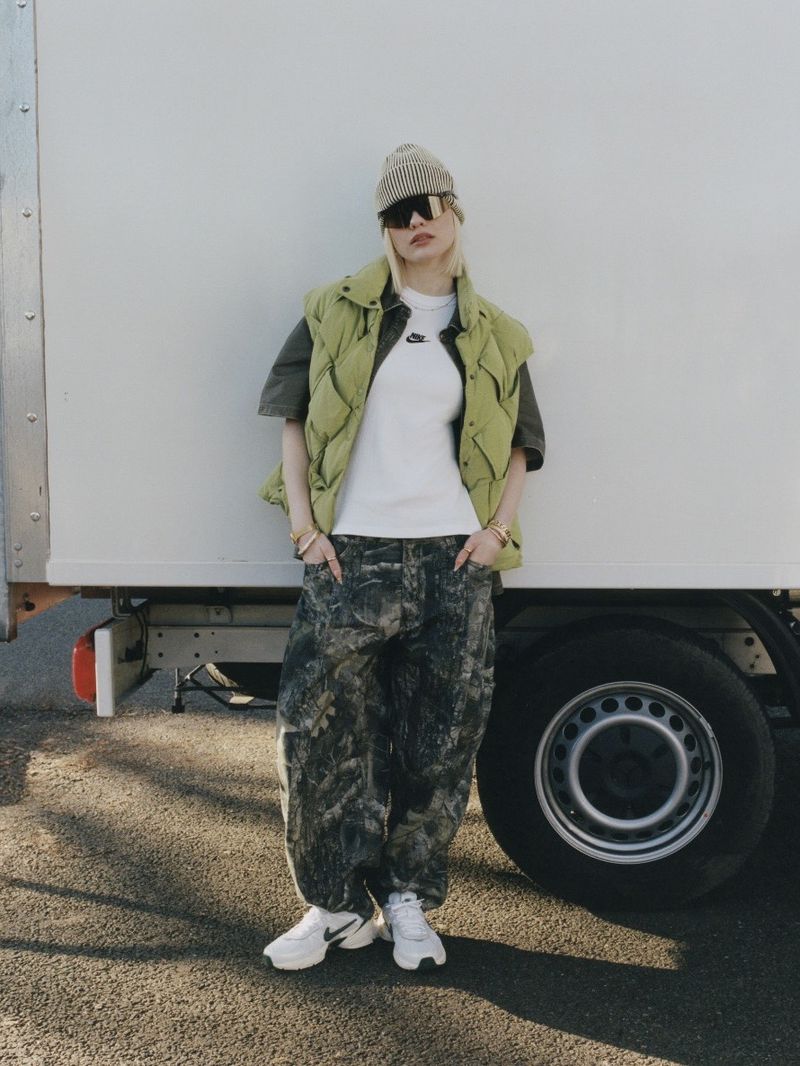 Woman in puffer vest, beanie, and baggy jeans stands in front of a truck, posing with hands in pockets, wearing sunglasses.