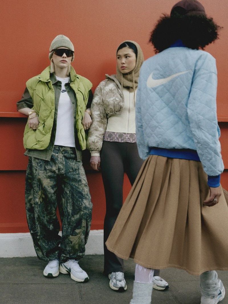 Three women wearing fashionable sportswear stand in front of a red wall in an urban setting, showcasing athletic apparel and urban style.