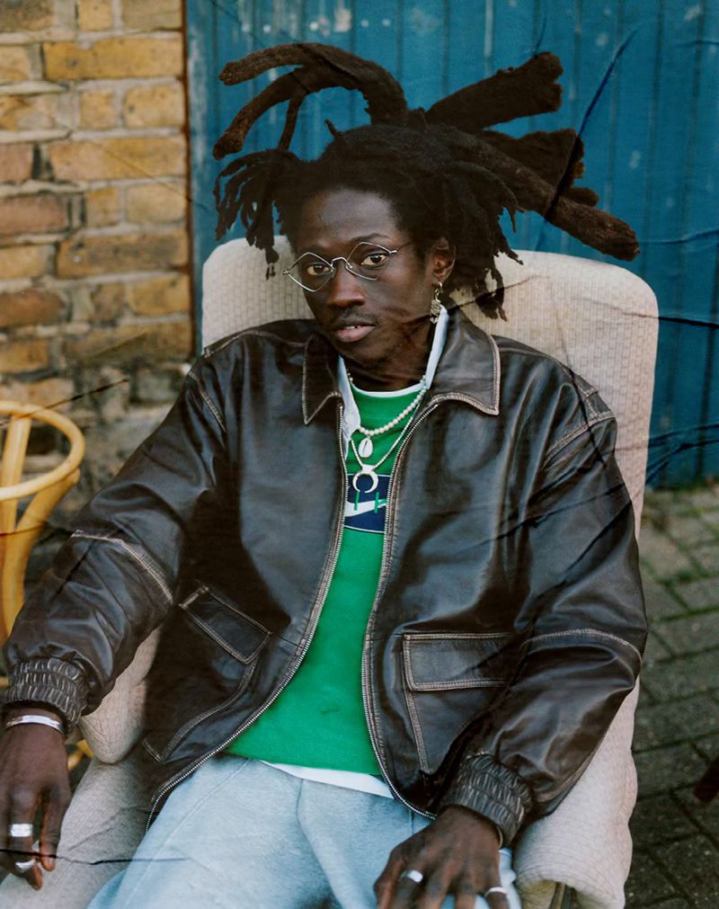 A black male model with dreadlocks is sitting in a chair outside wearing sunglasses, a green shirt, leather jacket, light pants, and jewelry.