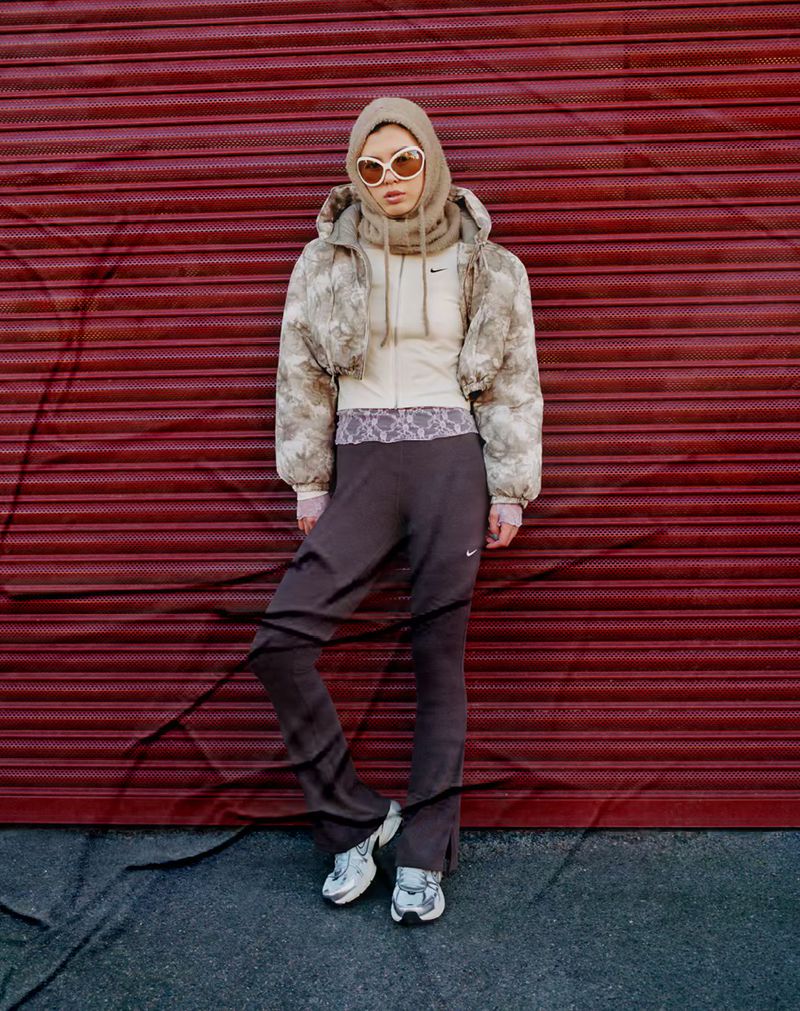 Full-length shot of a woman in athleisure wear with sunglasses, a cropped jacket, and running shoes standing against a burgundy metal door.