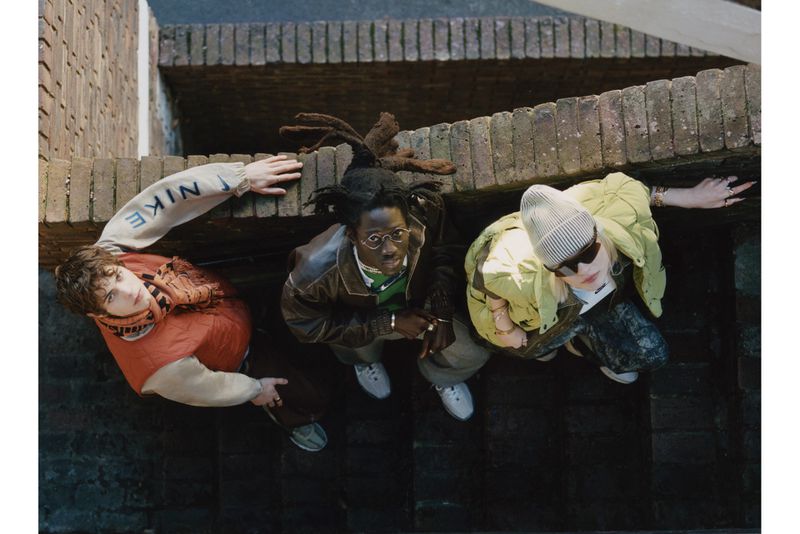 Three young adults in fashionable streetwear stand on steps in an urban outdoor environment, captured from an upward, eye-level angle.