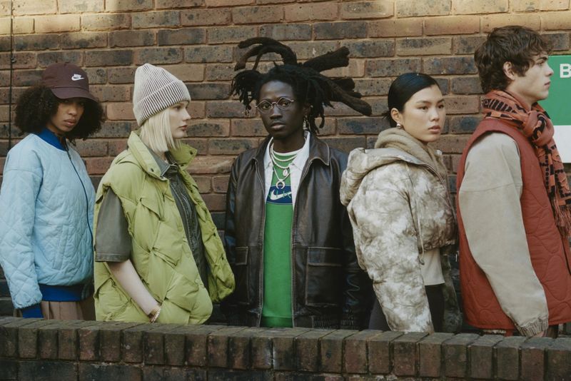 Group of five young adult models in jackets and casual wear stand against a textured brick wall for an outdoor fashion editorial.
