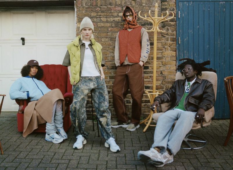 Four people model streetwear fashion against an outdoor brick wall, showcasing jackets, vests, pants, and sneakers in a casual setting.