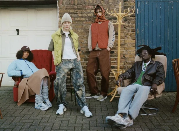 Four people model streetwear fashion against an outdoor brick wall, showcasing jackets, vests, pants, and sneakers in a casual setting.