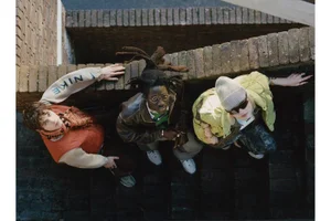 High-angle shot of three models in streetwear jackets and knitwear looking up from a brick outdoor stairway.