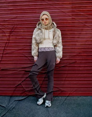 Model Maggie Yu in a beige balaclava, cropped puffer jacket, and brown track pants against a red corrugated shutter door.