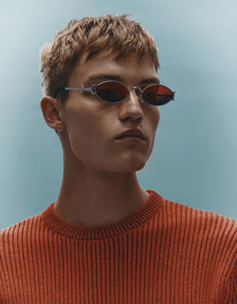 Close-up of a fair-skinned male model with red hair wearing oval sunglasses and an orange ribbed sweater against a light blue backdrop.