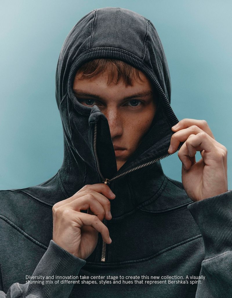 Close-up shot of a young adult male fashion model in a hooded sweatshirt with zipper detail, posing against a plain background in studio lighting.