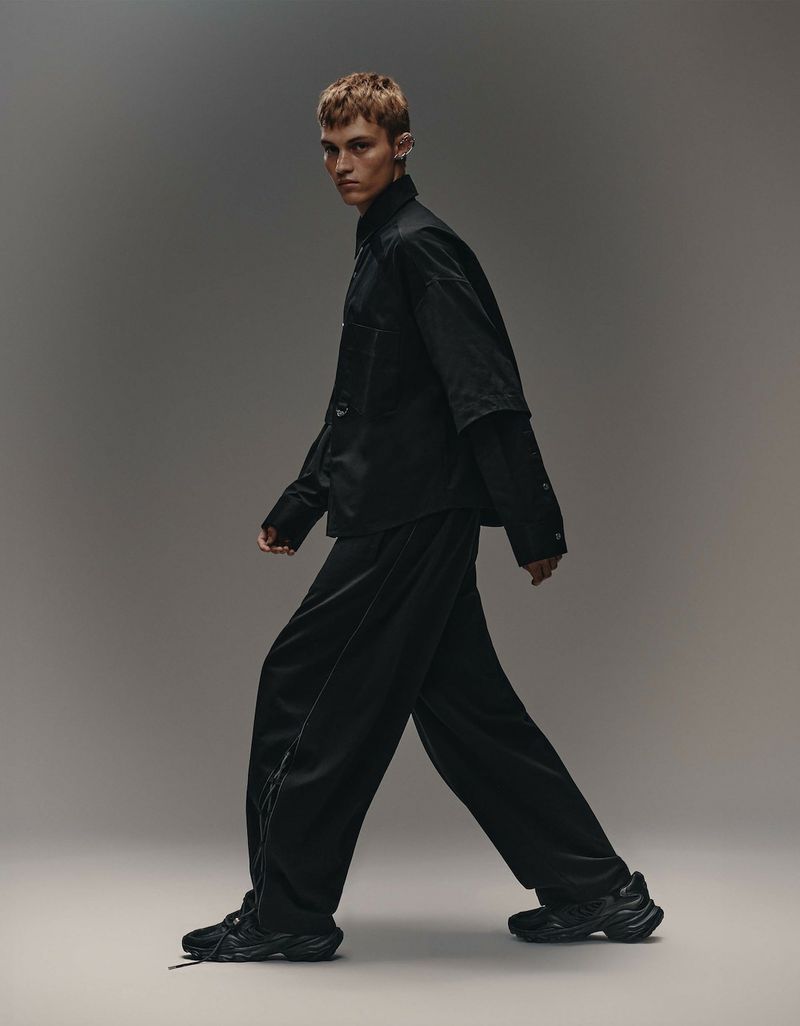 Full-length fashion portrait of a male model walking in a black jacket, black pants, and sneakers against a grey gradient background.