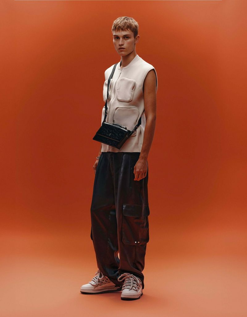 Full-length shot of a male model in a sleeveless top, cargo pants, and sneakers, with a black crossbody bag, against an orange backdrop.