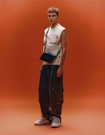 Full-length shot of a male model in a sleeveless top, cargo pants, and sneakers, with a black crossbody bag, against an orange backdrop.