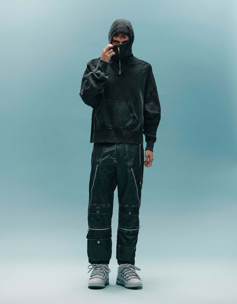 Full-body shot of a male model wearing a hooded sweatshirt, textured pants, and sneakers in a studio with light blue background.