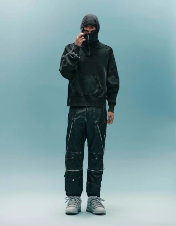 Full-body shot of a male model wearing a hooded sweatshirt, textured pants, and sneakers in a studio with light blue background.
