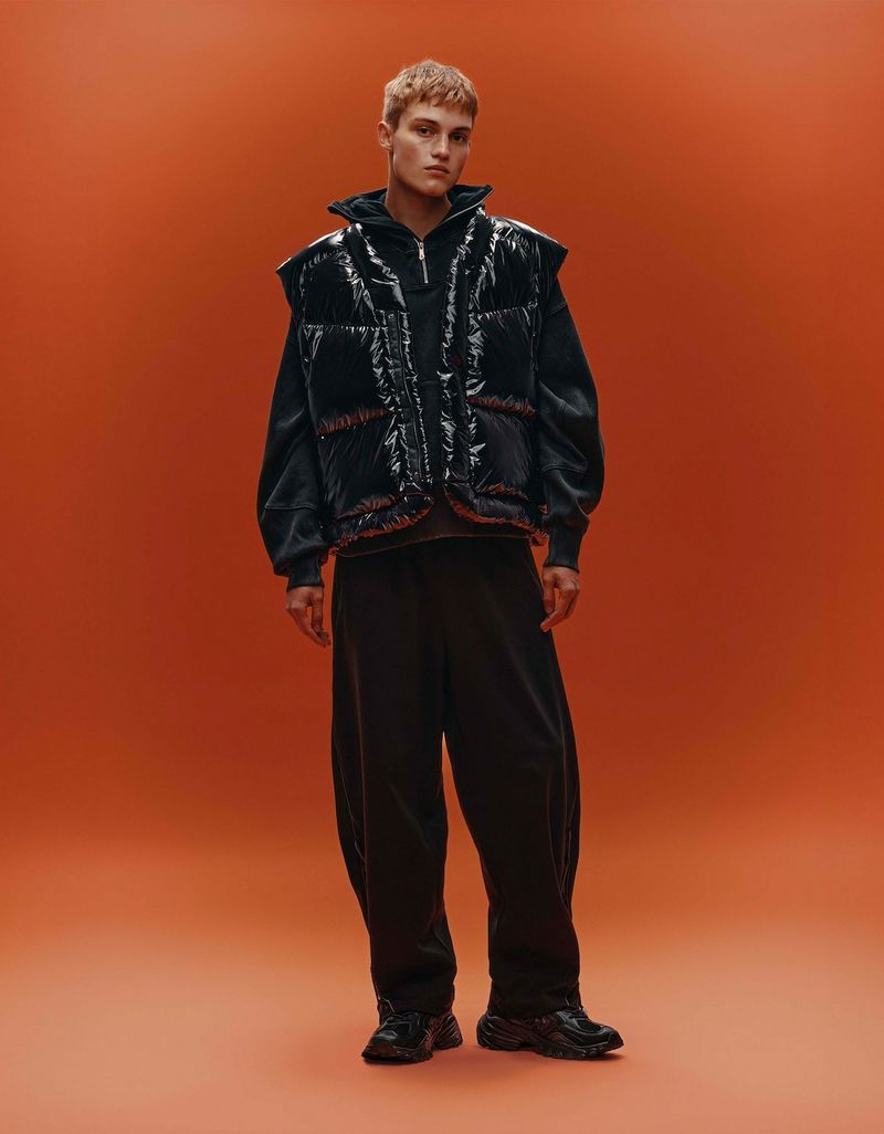 Model Dominik Datko wearing a black glossy puffer vest and wide-leg trousers against a solid orange studio backdrop.