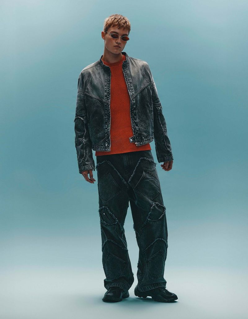 Fashion model poses in distressed denim jacket and loose pants against a soft blue backdrop, showcasing a relaxed, modern style.