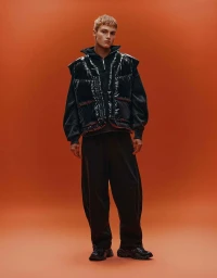 Model Dominik Datko wearing a black glossy puffer vest and wide-leg trousers against a solid orange studio backdrop.