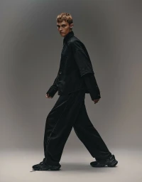 Dominik Datko walking in an all-black layered shirt, wide-leg trousers, and chunky sneakers against a grey studio backdrop.