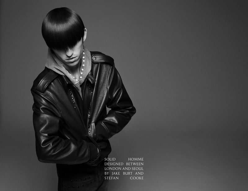 Black and white studio shot of a young adult man wearing a black leather jacket, hoodie, and dark pants, with dark hair.