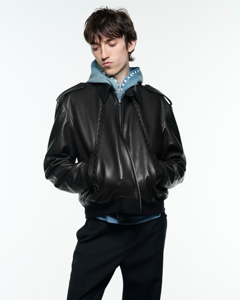 Adult male fashion model poses in a black leather jacket, blue hoodie, and black pants against a white studio backdrop.