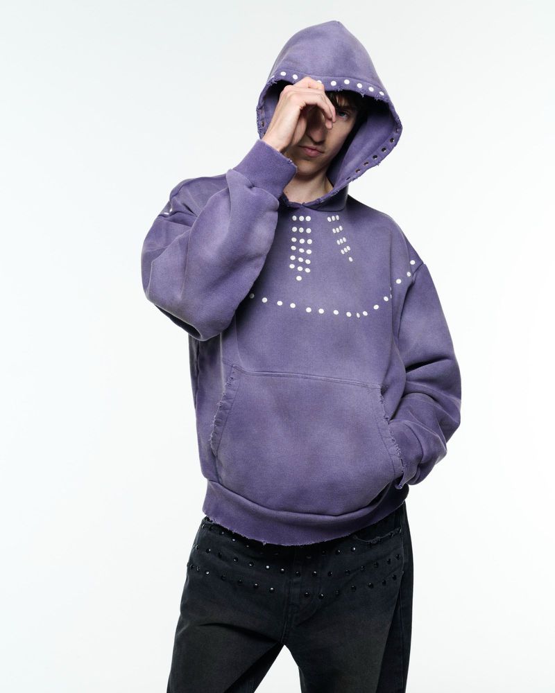 Adult male with his hand near his face, wearing a faded purple studded hoodie and black studded jeans in a bright, neutral studio.