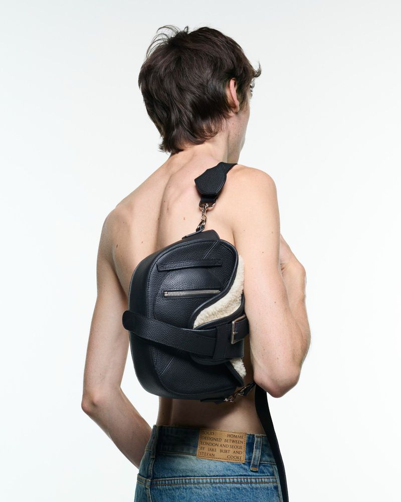 An adult man is seen from the rear, wearing light blue jeans and carrying a black leather and faux fur bag over his shoulder.