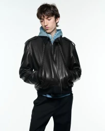 A male model wearing a black leather bomber jacket over a light blue studded hoodie against a pure white studio backdrop.
