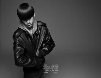 A young male model with a heavy bowl haircut wearing a black leather jacket and grey hoodie against a plain grey backdrop.