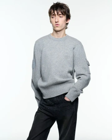 Model wearing a grey knit crewneck sweater and studded black jeans against a white seamless studio backdrop.
