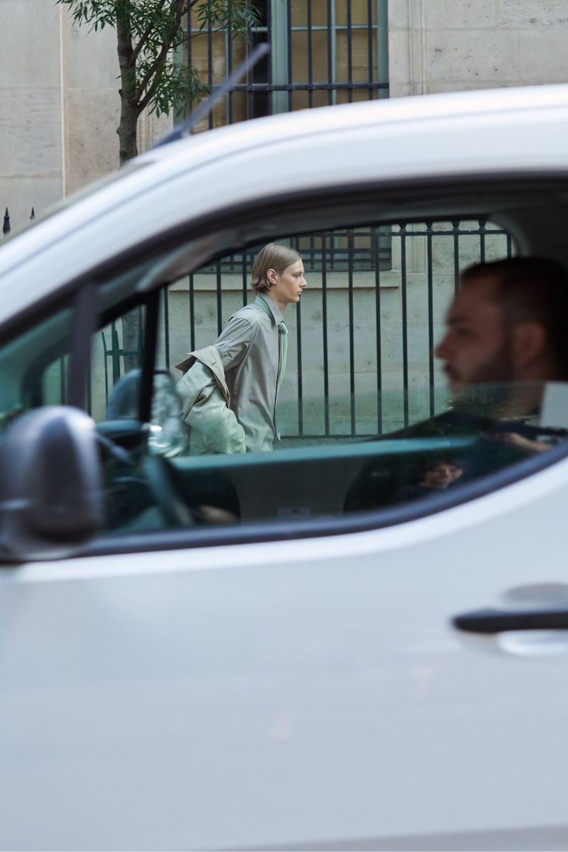 A fashion shot framed through a vehicle window, featuring a male model in a light outfit walking near a building with a metal fence.