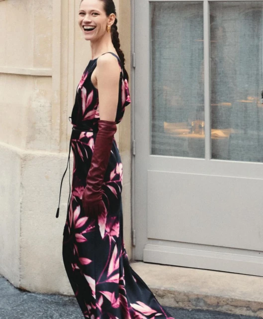 Smiling woman in a floral dress and long gloves standing outside near a door. The background is a light beige wall and sidewalk.