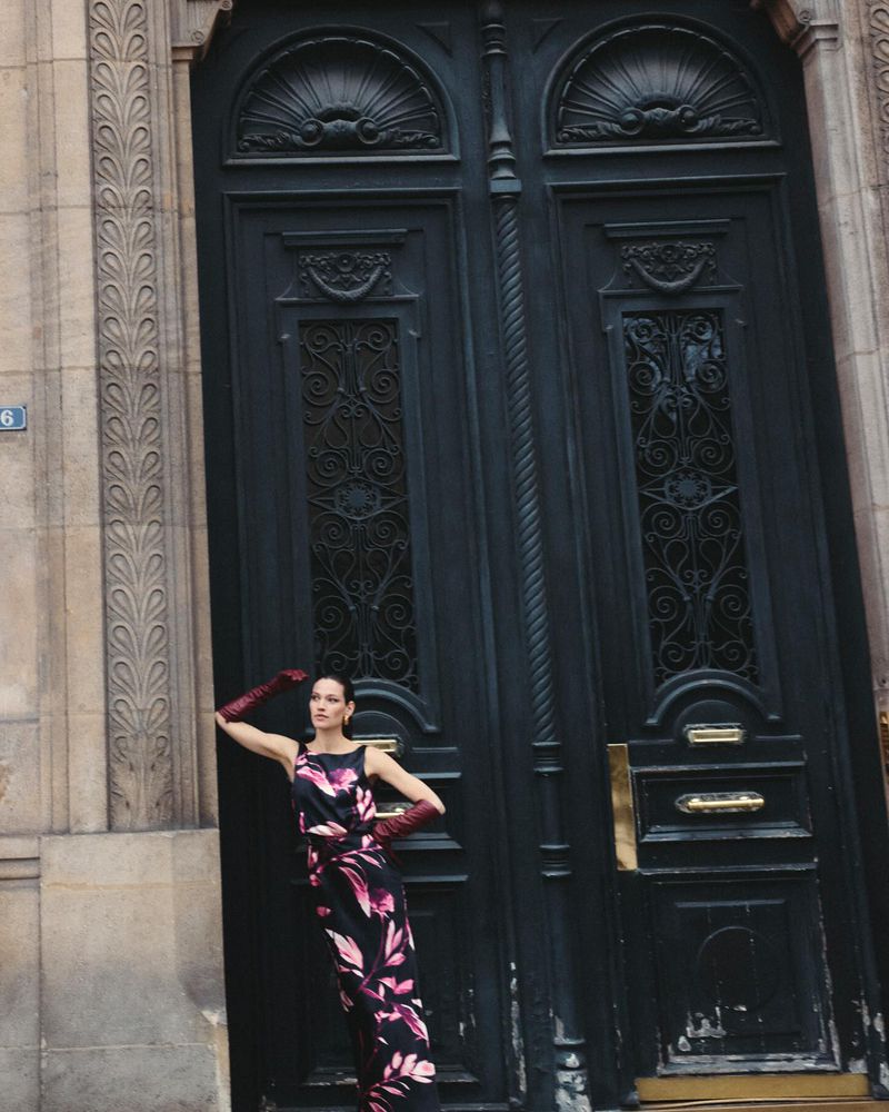 A woman in a floral dress poses in front of a large, ornate, dark gray door in an outdoor urban environment with vintage architecture.