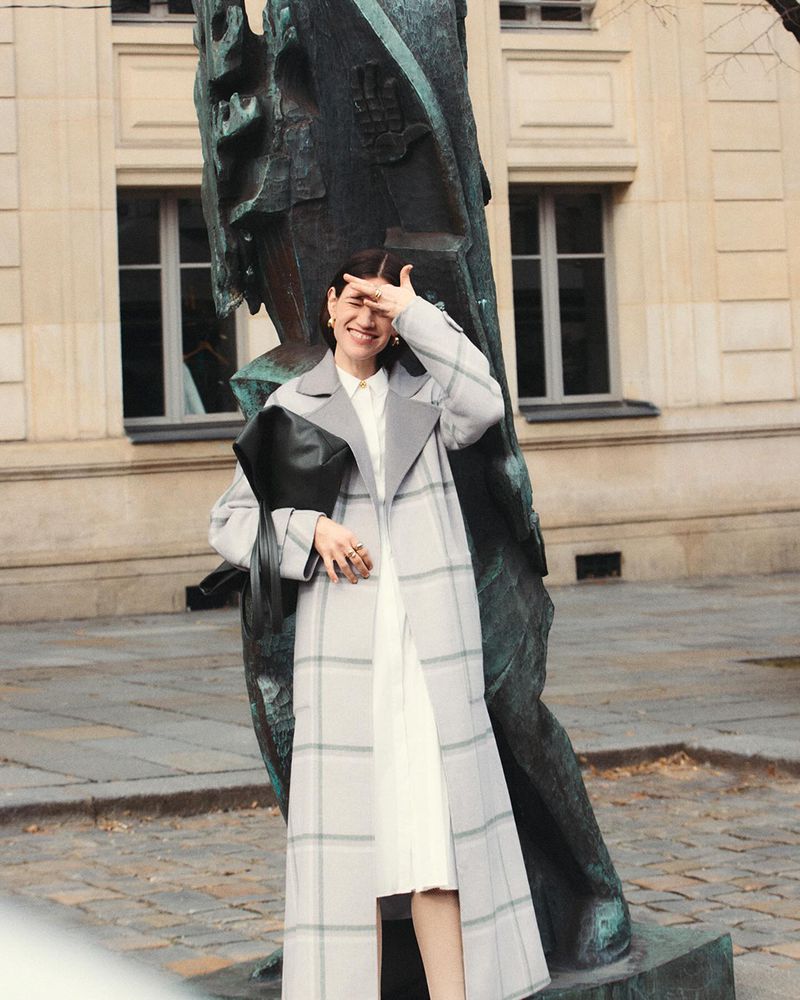 A smiling woman in a gray plaid coat poses next to a statue with a black clutch, outdoors with a building in the background.