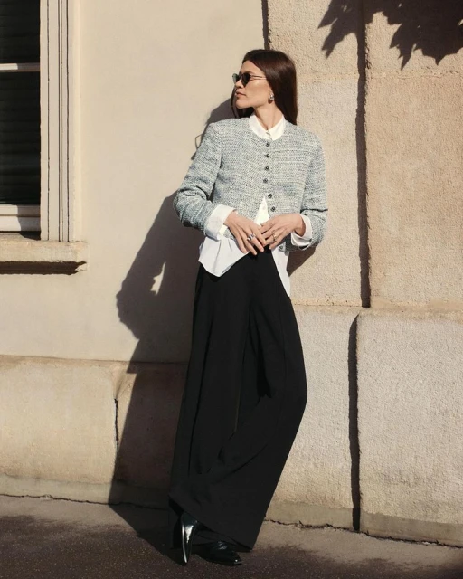 Fashionable woman in blazer, blouse, and palazzo pants poses against wall with tree shadow in sunlight, accessorized with sunglasses.