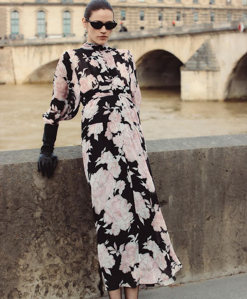 Adult woman in a floral dress and gloves poses on a bridge with arches in the background. Fashion shot with a slightly overcast day.