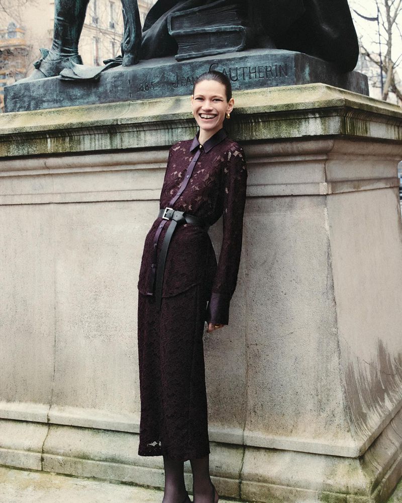 A woman in a maroon lace dress with a belt stands in front of a stone monument smiling in an outdoor urban setting during daytime.