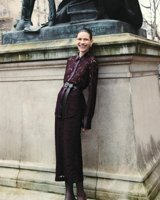 A woman in a maroon lace dress with a belt stands in front of a stone monument smiling in an outdoor urban setting during daytime.
