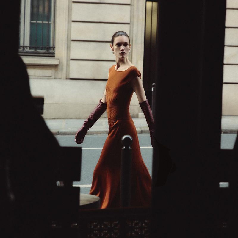 An adult woman wearing a long copper-colored dress and burgundy gloves walks on a city sidewalk near a building with windows.