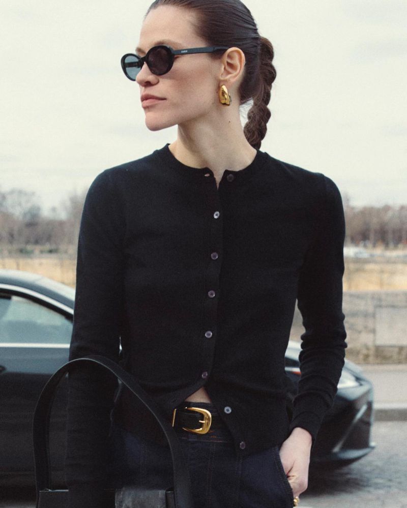 Adult woman in black cardigan, sunglasses, and gold earrings stands outside next to a luxury car with cloudy sky.